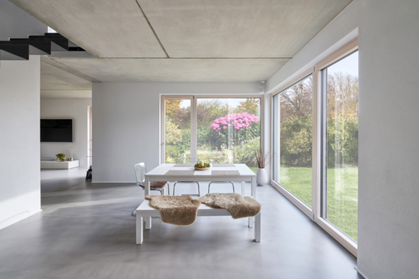 Blick in einen Wohnraum mit raumhoher Übereck-Verglasung und einem im Raum stehenden weißen Esstisch auf geschliffenem Betonboden