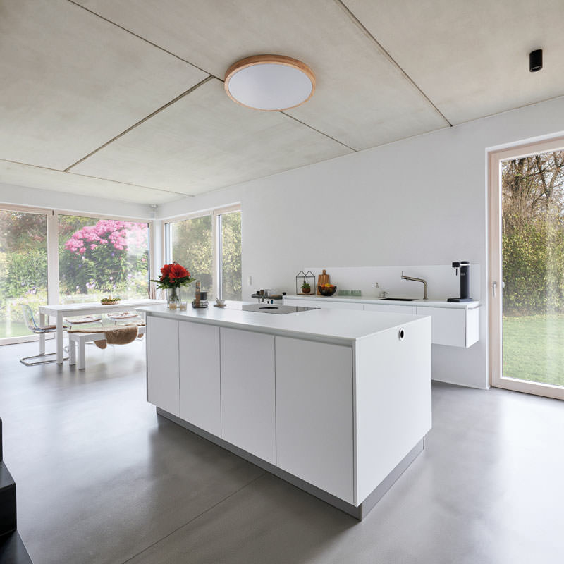 Ein weißer Küchenblock steht auf blank poliertem Betonfußboden, im Hintergrund ein weißer Esstisch, links im Bild führt eine schwarze Treppe in OG.