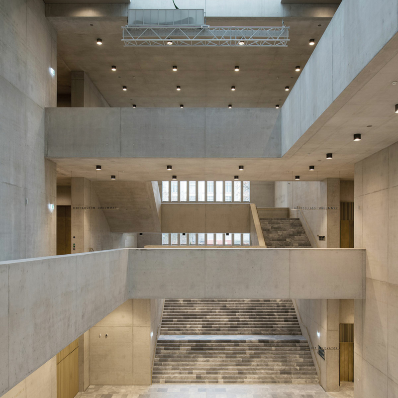 Halle mit grauem Steinboden, breitem Treppenaufgang mit grau marmorierten Stufen, Galerien in Sichtbeton, einer bodentiefen gerasterten Fensterfront auf der gegenüberliegenden Seite und Oberlicht
