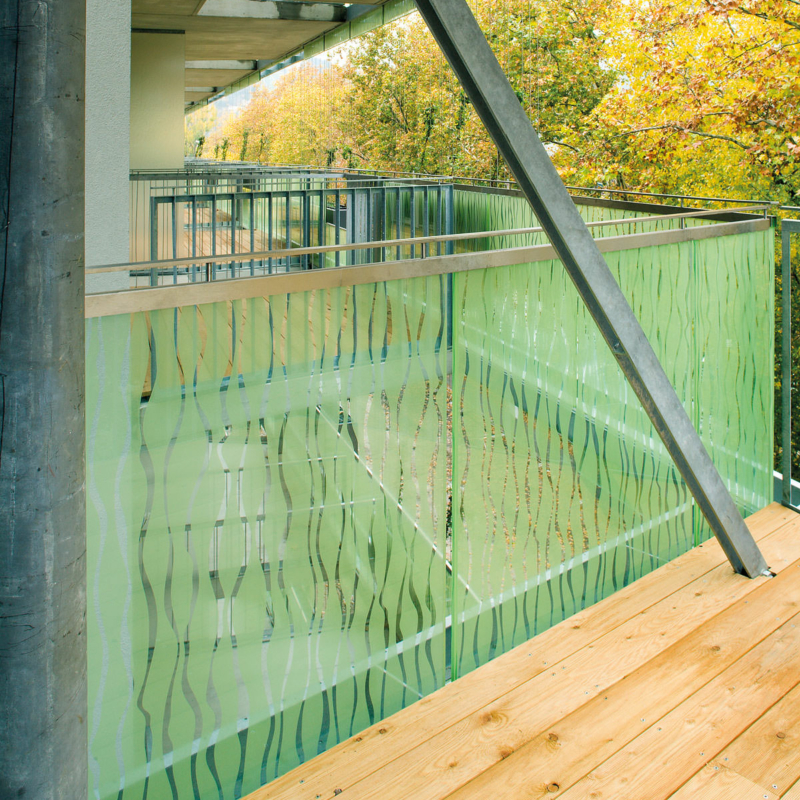 Teil eines Balkons mit dezent grün mit unregelmäßigem Liniendekor bedrucktem Glasgeländer, davor heller Holzfußboden und eine schräg montierte Metallstütze