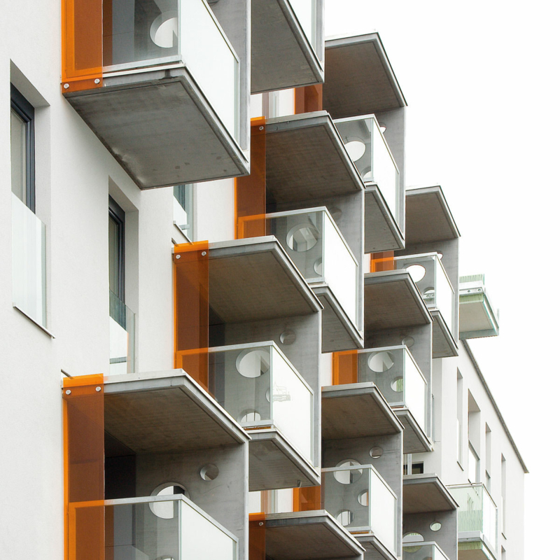 Eine weiße Fassade mit vorgesetzten, an einer Seite geschlossenen Balkonen, an deren anderen Seiten orangefarbene Gläser angebracht sind.