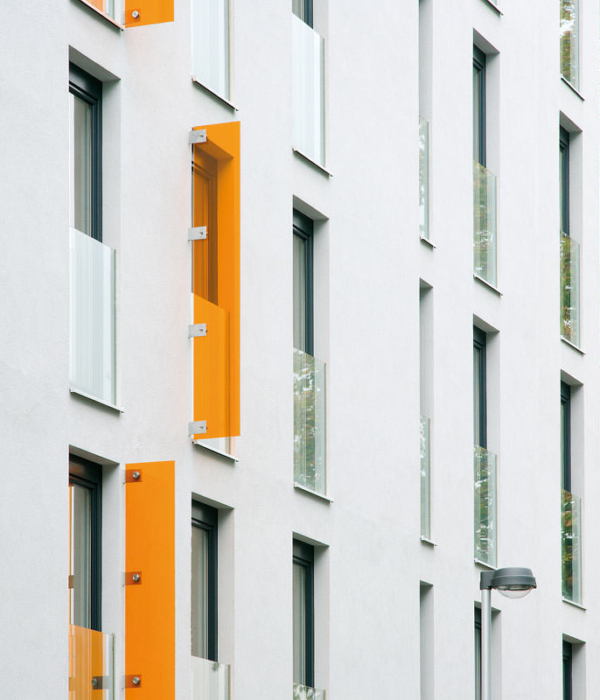Hohe schmale Fensteröffnungen mit vorgesetzten halbhohen Glasscheiben in einer großen weißen Fassadenfront. Einige schmale orangefarbene Gläser sind dazwischengesetzt.