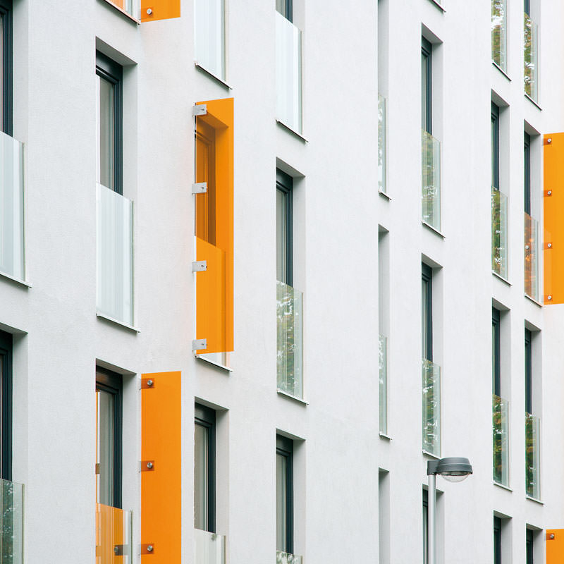 Hohe schmale Fensteröffnungen mit vorgesetzten halbhohen Glasscheiben in einer großen weißen Fassadenfront. Einige schmale orangefarbene Gläser sind dazwischengesetzt.