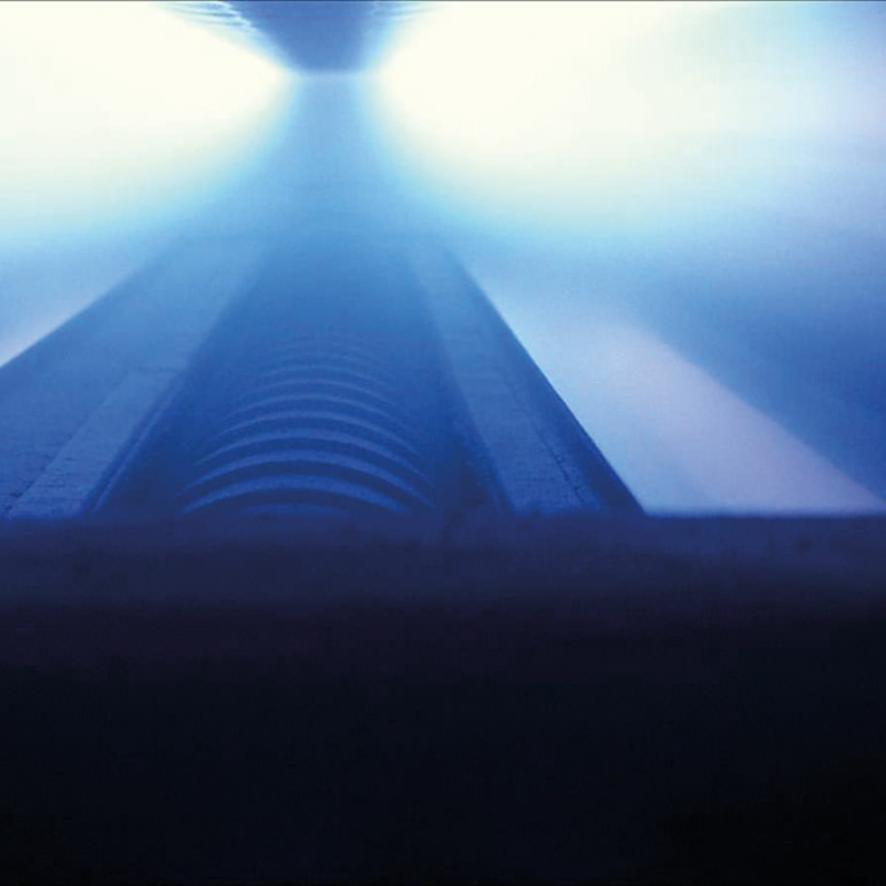 Das Innere einer Magnetron-Anlage, unscharf eine Walze und Sprühnebel in Blautönen, von dunkelblau vorne bis fast weiß wie überbelichtet hinten.