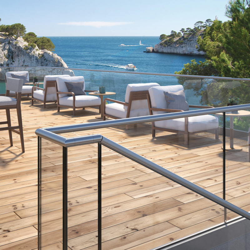 Ein Glasgeländer mit Edelstahlhandlauf umgibt eine große Terrasse mit niedrigen Sitzmöbeln und Holzboden vor dem Panorama einer felsigen Meeresbucht.