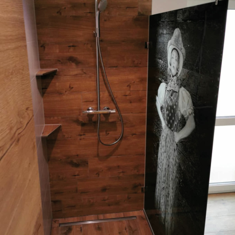 Dusche mit einer gläsernen Abtrennung in schwarz, auf die das Motiv eines Schwarzwaldmädels gedruckt ist.