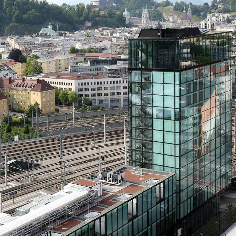 Gebäude mit gläsernem Turm vor Bahngleisen und der Altstadt von Salzburg
