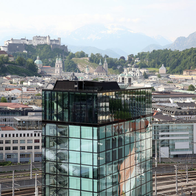 Gläserner Turm vor Bahngleisen und der Altstadt von Salzburg
