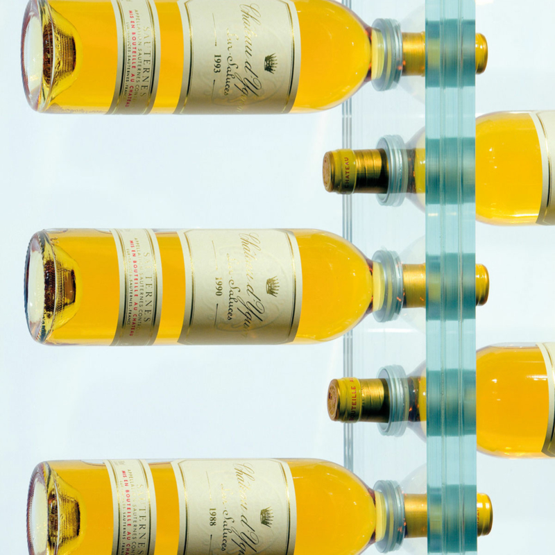 In einer aus drei Scheiben bestehenden schmalen, längs von oben nach unten reichenden Glasplatte, deren Schnittkanten zu sehen sind, stecken links und rechts abwechselnd Weinflaschen in regelmäßig in der Glasplatte angebrachten Öffnungen.
