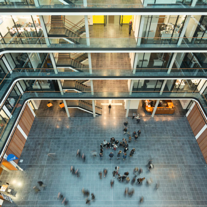 Blick vom sechsten Stock in ein großes Atrium eines Bürogebäudes; im EG halten sich mehrere Personen auf.