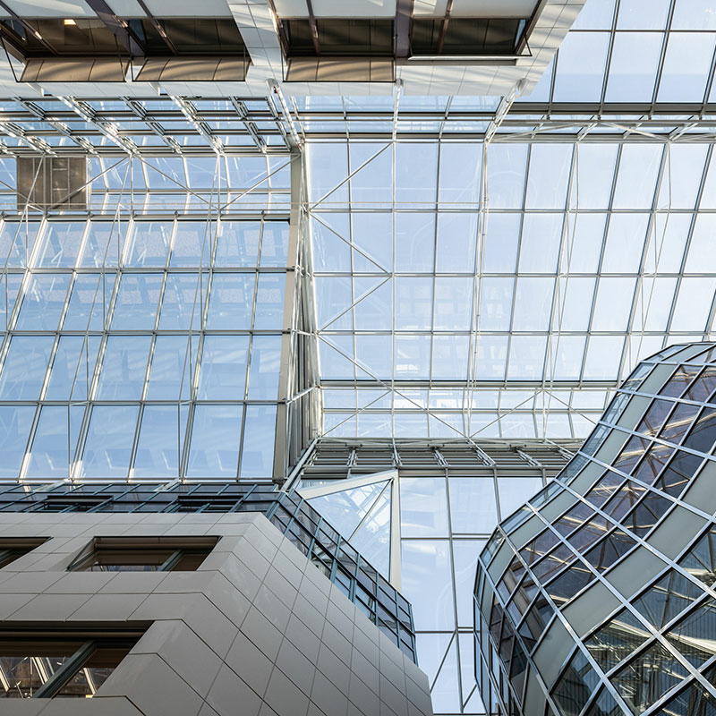 Blick von unten durch ein Glasdach mit schlanken, hellen Rahmenprofilen in blauen Himmel. Links unten Teil einer mit rechteckigen, hellgrauen Platten verkleideten Fassade, rechts ein Teil einer gewellten Glasfassade.