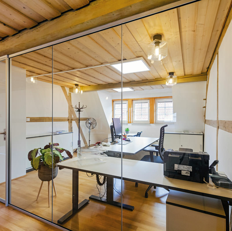 Blick in einem Dachgeschoss mit Holzbalken durch eine gläserne Wand mit Tür in einen Arbeitsraum