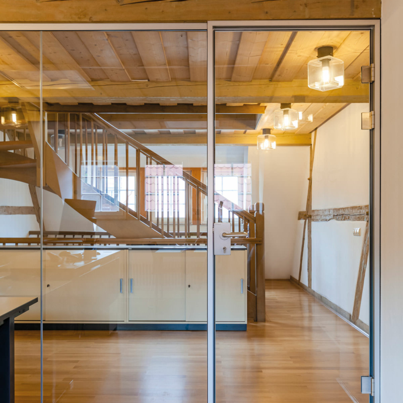 Teil einer Ganzglaswand mit Glastür in einem Fachwerkgebäude, dahinter ein Flur mit Treppen