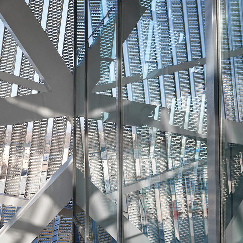 Detail der Metalllamellenkonstruktion von innen: links im Bild sechs Lammellenbänder mit gleichmäßigem Abstand, rechts eine Glaswand, dahinter sich überlagernde Lamellen durch Spiegelungen im Glas.