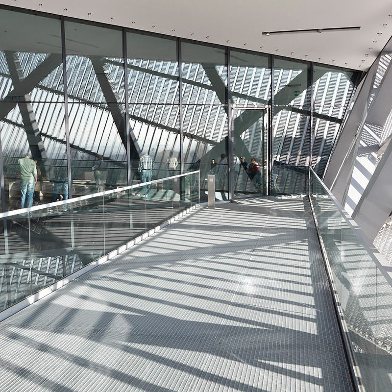 Eine Metallbrücke im Innern des Keils, mittig im Bild. Glasgeländer links und rechts, mit hellem Vier-Kant-Handlauf. Die Brücke läuft auf eine, durch schlanke, dunkelgraue Profile unterteilte Glasfassade zu, dahinter sieht man die Metallstruktur, durch diese wiederum kann man den Himmel erahnen.