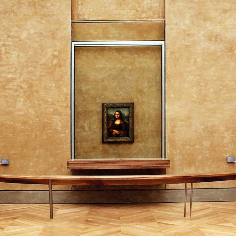 An einer gold-/ockerfarben wie gewischt aussehenden Wand hängt das gerahmte Gemälde der Mona Lisa, umgeben von einem sehr großen Glasrahmen, den unten ein dickes Holzbrett abschließt. Unter dem Bild, auf einem Fußboden aus hellem Holzparkett, steht eine leicht nach unten durchgebogene Holzbank.