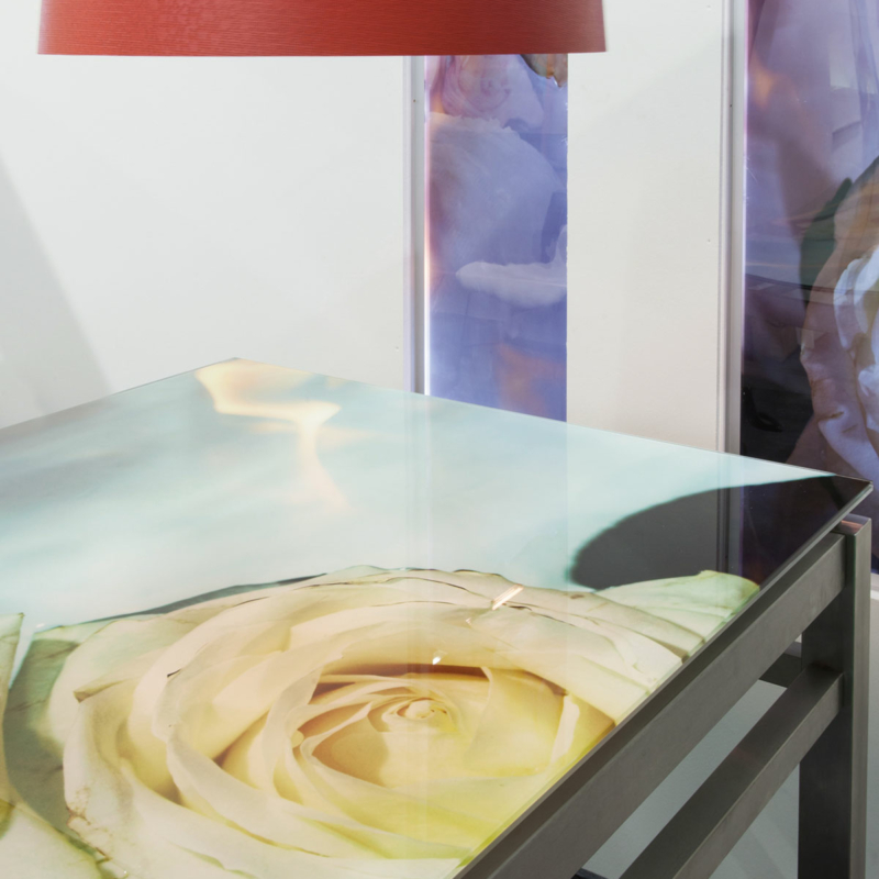 Tisch mit einer gläsernen, mit einem großen Rosenmotiv bedruckten Platte auf einem Metallgestell. Darüber ein roter, runder Lampenschirm und im Hintergrund in einer weißen Wand zwei schmale, mit floralen Motiven in Lila- und Rosatönen bedruckte Glaspaneelen.