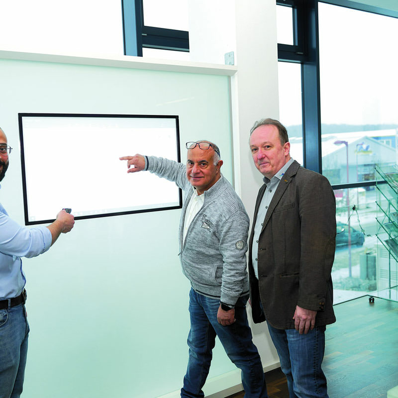 Drei Männer stehen in einem Raum mit bodentiefer Verglasung vor einem Display mit eingelassenem Bildschirm.