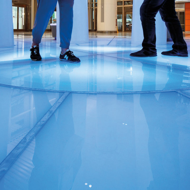 Auf einem blauen Glasfußboden sind Füße und kniehoch Beine von zwei Personen zu sehen. Im Hintergrund begrenzen weiße Säulen und Glastüren die Fläche.