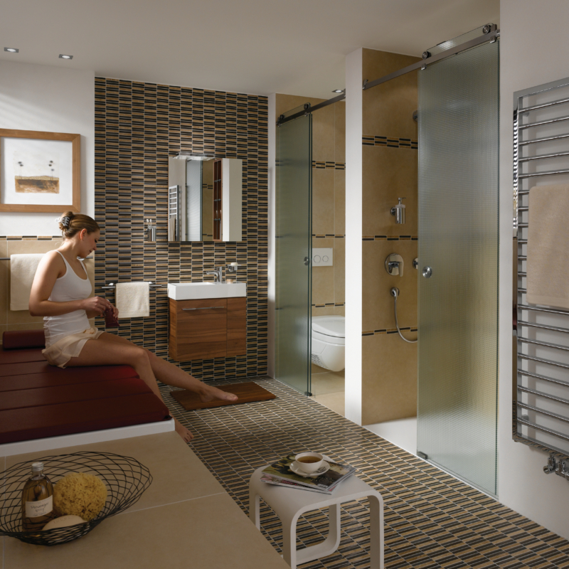 Badezimmer in Beigebrauntönen. Auf der rechten Seite sind eine Dusche und ein WC durch strukturierte Ganzglasschiebetüren abgetrennt. Links sitzt eine Frau auf einem braunen Leder- oder Holzpodest.