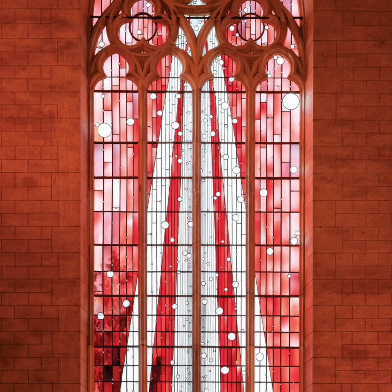 Schmales hohes Kirchenfenster mit vielen kleinen Gläsern in unterschiedlichen Rottönen.