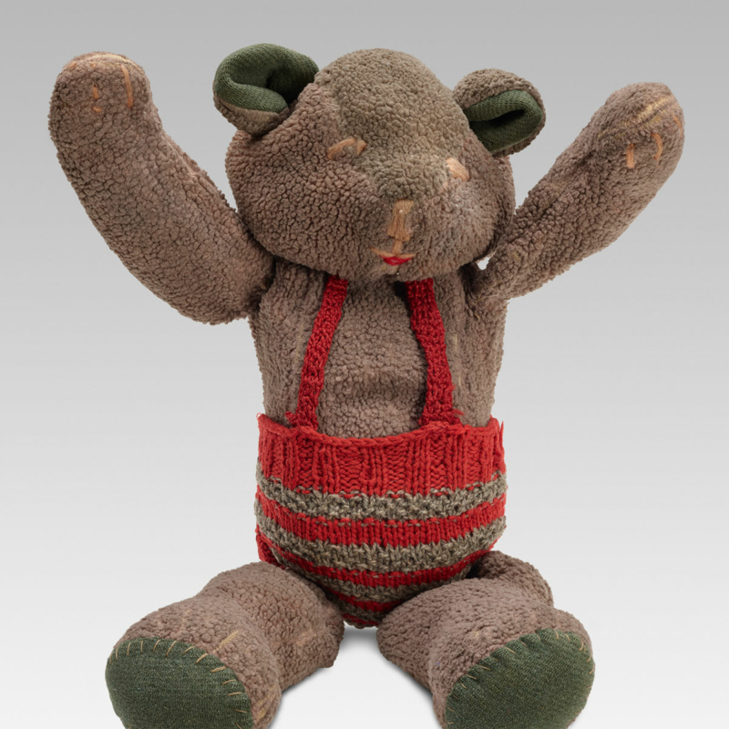 Ein graubrauner kleiner Stoff-Teddy mit rot-grau gestreifter, gestrickter Hose mit roten Hosenträgern. Die Beine zeigen nach vorne, als ob der Teddybär sitzen würde, die Arme sind nach oben gestreckt.