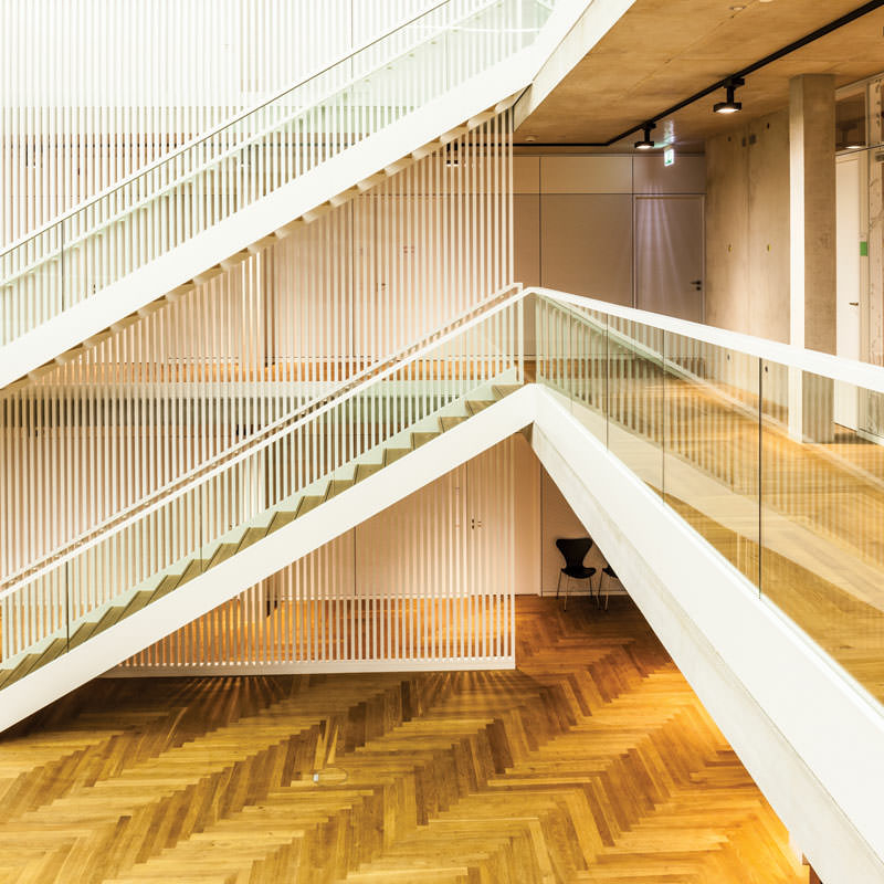 Zwei schräge parallel verlaufende Treppen mit Glasgeländern vor einem weißen gestreiften (Vorhang-)Element in einer großen Halle mit Holzfußboden in Frontalansicht. Rechts eine Galerie mit Holzfußboden und Glasgeländer vor großen Glastüren.