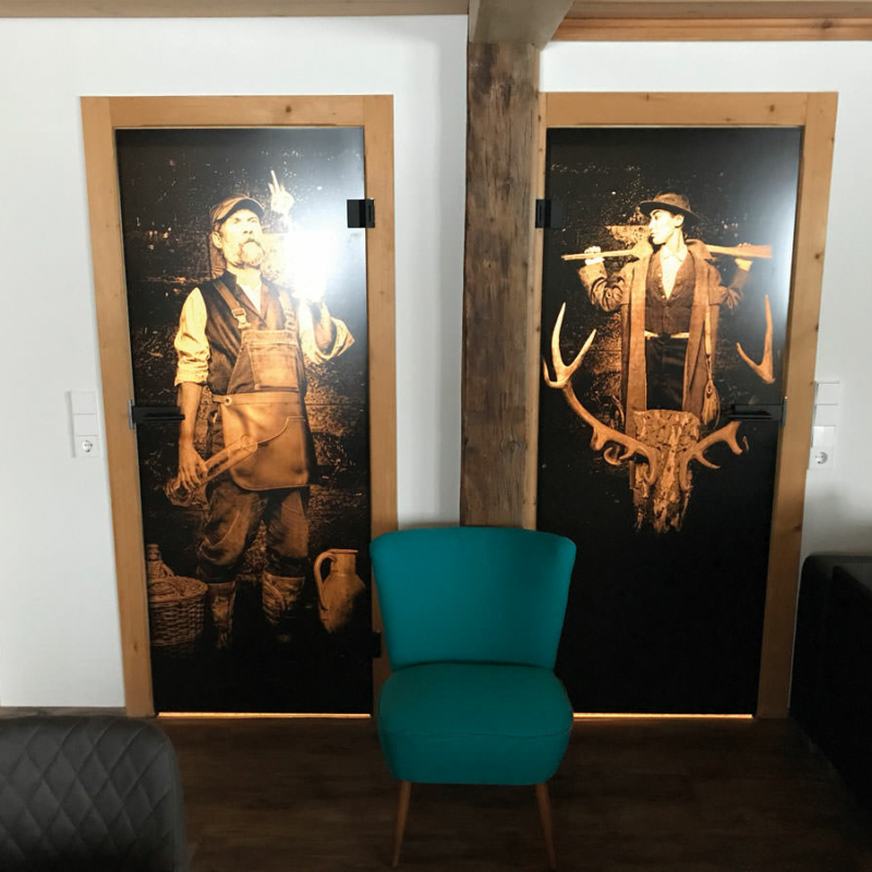 Ein Raum mit zwei schwarz beschichteten Ganzglastüren bedruckt, einmal mit einem Foto eines Schnapsbrenners und einmal mit dem Motiv “Jagd” bedruckt.