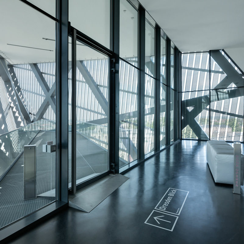 Blick durch große Glastüren auf eine kleine Aussichtsplattform vor einer Fassadenverkleidung aus schmalen Stahlstreben; auf dem Boden zeigen ein Pfeil und die Wörter „Dresden Bild“ die Richtung zur Aussichtsplattform an.