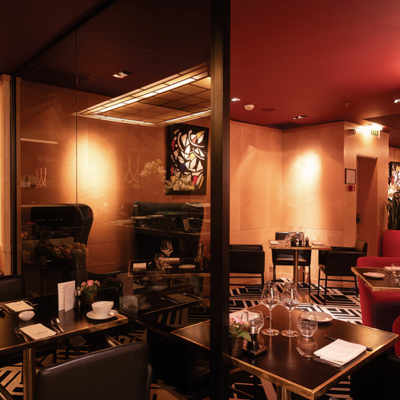 Ein in dezenten Rot-, Beige- und Brautönen gehaltener Restaurantbereich in einem Hotel. Eine transparente bronzefarbene Ganzglaswand trennt einen Bereich teilweise ab.