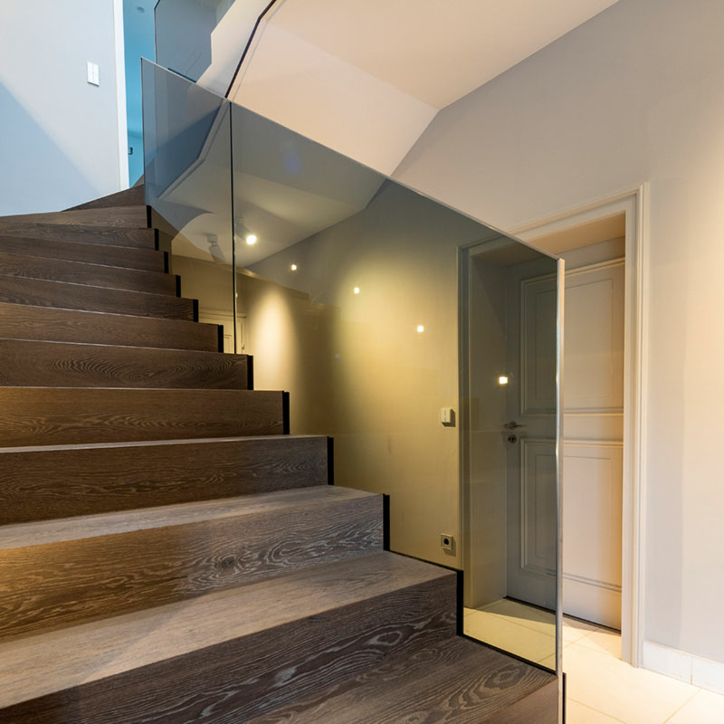 Treppe mit dunklen Holzstufen, die sich mit leichter Rechtsdrehung zu einem Treppenabsatz hochzieht in einem Wohnhausflur mit weißen Wänden, Tür und Boden. Die Treppe ist von einem Ganzglasgeländer aus transparent-grauem Glas begrenzt.