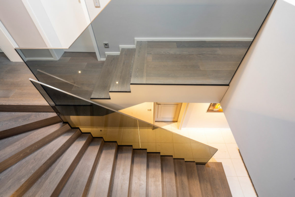 Blick von oben in einen Hausflur mit Treppenhaus mit dunklen Holzstufen. Ein zweigeteiltes Ganzglasgeländer aus transparent grauem Glas begrenzt die Stufen und schließt oben an einer weißen Wand ab.