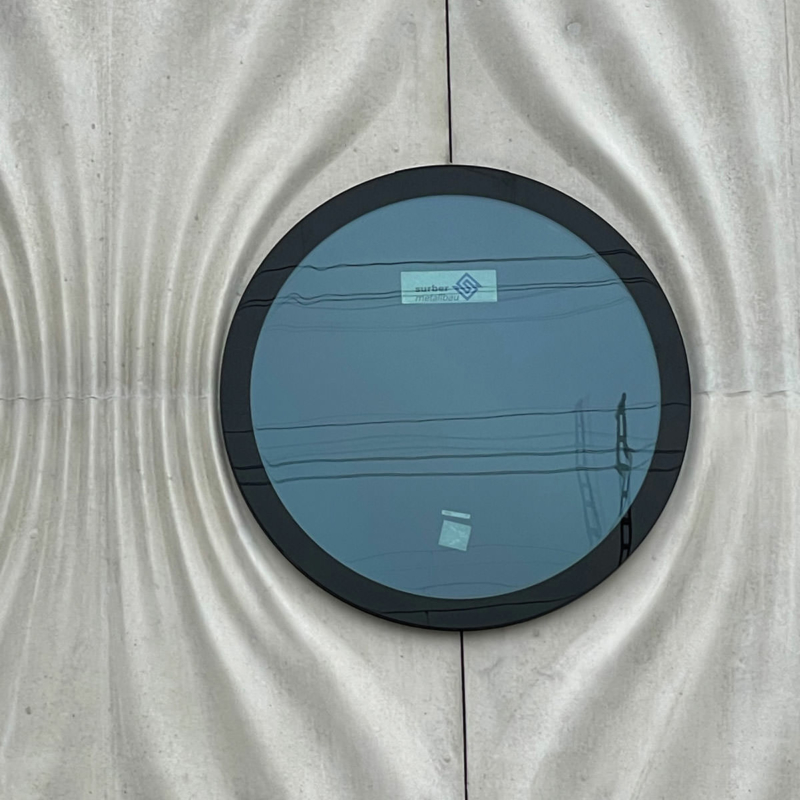 Nahaufnahme eines runden Fensters, das bündig zur gefalteten Betonstruktur der umgebenden weißen Fassade montiert ist. schwarzer Siebdruck im Randbereich verdeckt den Glasverbund.