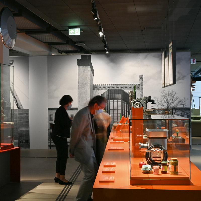 Ausstellungsraum mit verschiedenen Glasboxen auf orangefarbenem Grund. Mittig beugt ein Mann mit unscharfem Gesicht die Beine, um sich eine Ausstellungsvitrine genauer anzuschauen.