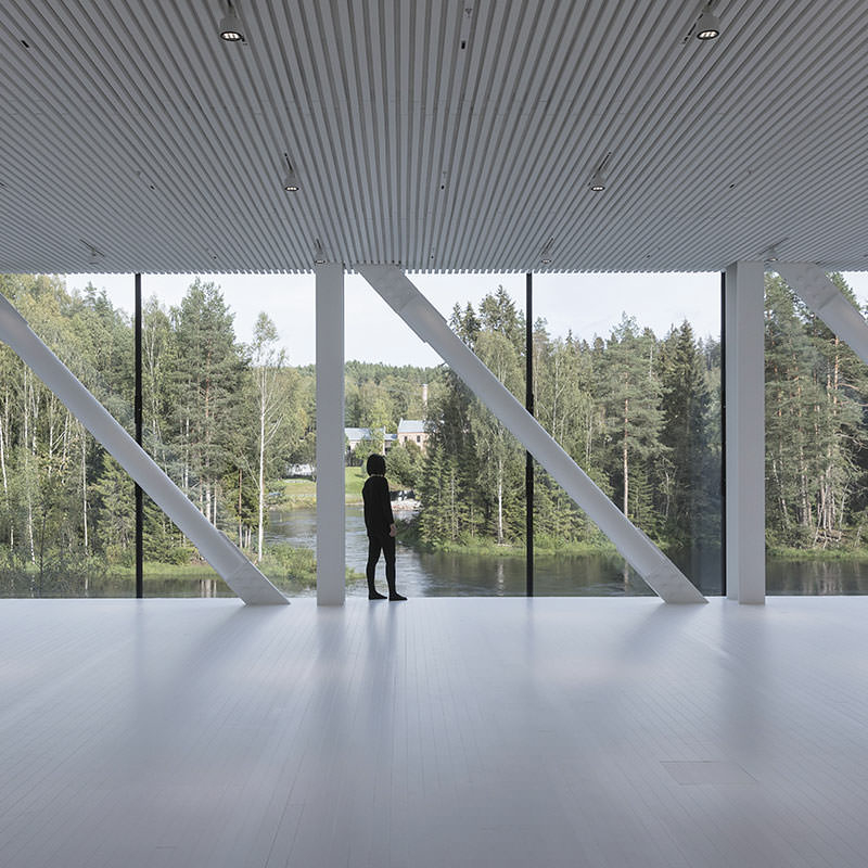 Blick durch Panoramafenster einer Halle mit weißem Boden und weißer Decke in den Wald