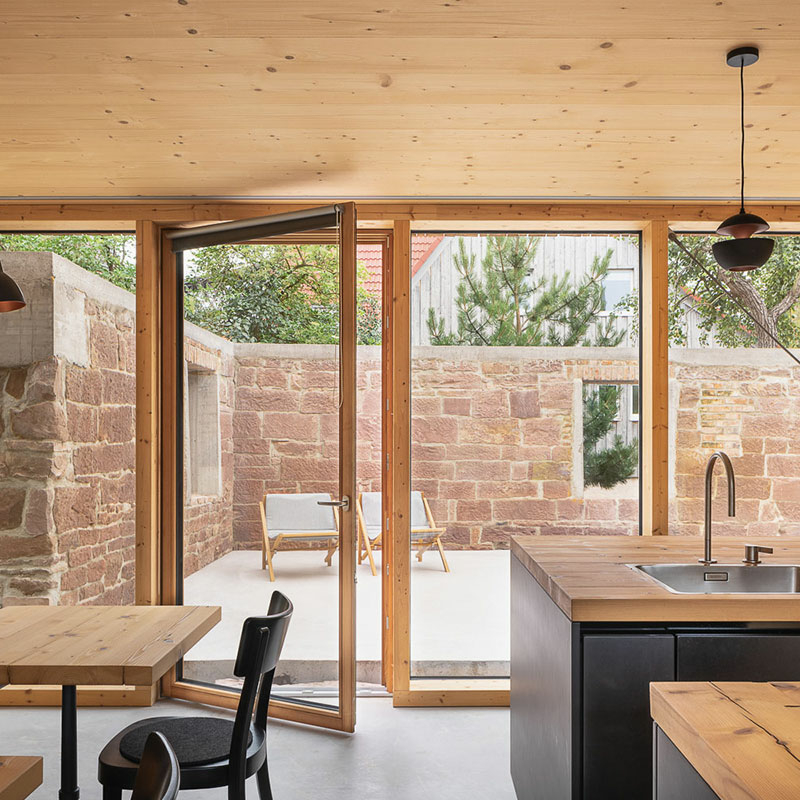 Ein Wohnzimmer mit gläserner Doppelflügel-Terrassentür, durch die man in einen Hof blickt, der mit einer Mauer aus großen Natursteinquadern umgeben ist. Rechts ein Küchentresen mit Holzoberfläche und Spülbecken.