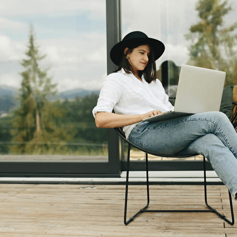 Eine junge Frau, bekleidet mit Jeans, weißer Bluse und schwarzem Hut und barfuß, sitzt lässig auf einem Stuhl, mit einem Laptop auf dem Schoß, vor einer großen, zweiteiligen, bodentiefen Glasschiebetür