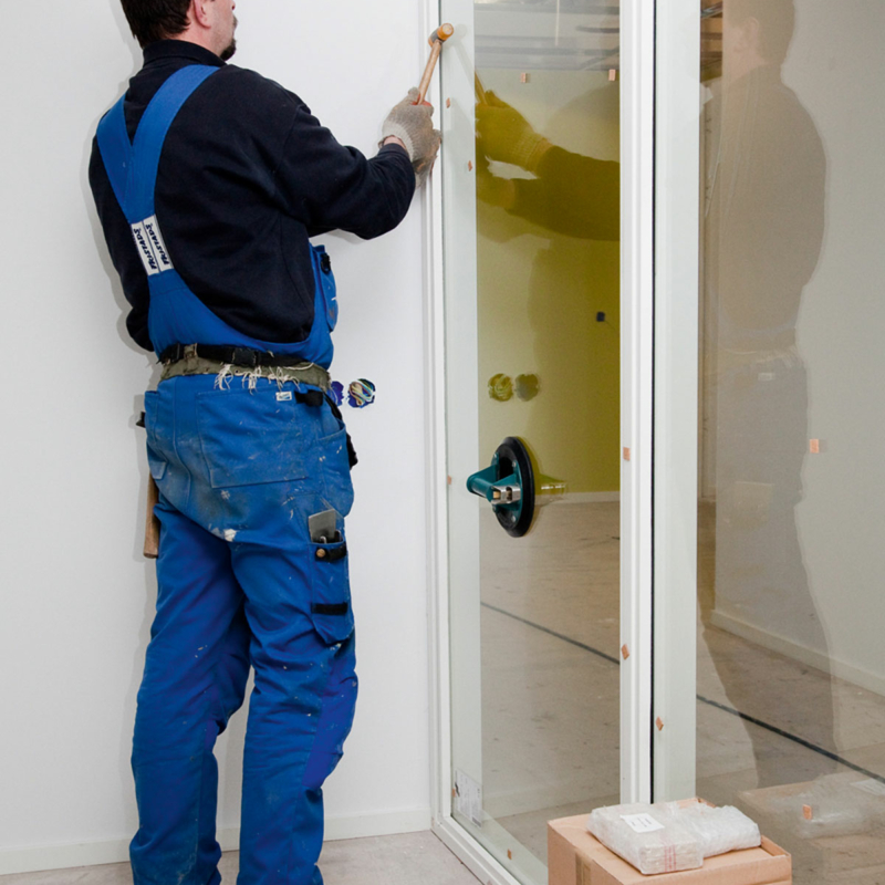 Ein Mann in blauem Overall und schwarzem Pulli kniet vor einem Glaselement in weißem Rahmen, in der rechten Hand hält er einen kleinen Gummihammer. Rechts von ihm steht an eine weiße Tür gelehnt eine neue Glasscheibe ohne Rahmen.