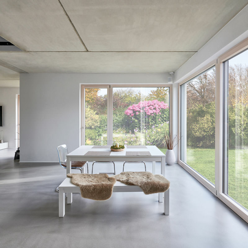 Blick in einen Wohnraum mit raumhoher Übereck-Verglasung und einem im Raum stehenden weißen Esstisch auf geschliffenem Betonboden|Wohnbereich des EFH im EG