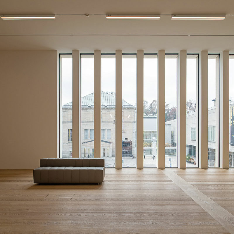 Großer Saal mit einer großen durch längs angeordnete Balken unterteilten Fensterfront und Holzboden