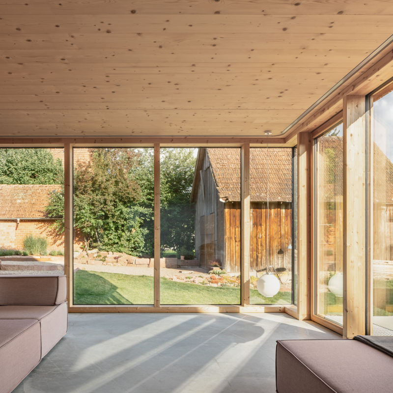 Über-Eck-Blick durch bodentiefe Fenster mit Holzrahmen nach draußen auf eine Holzscheune und Rasen
