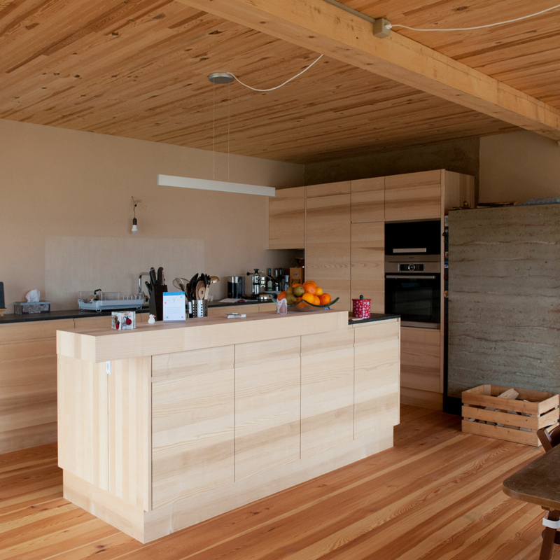 Großer Wohnraum mit zentralem Küchenblock. Decke, Boden und Möbel sind aus Holz in unterschiedlichen Farbtönen. Rechts im Bild ein dunkelgrauer Lehmofen, links eine offene Tür, die den Blick auf eine graue Lehmwand mit schmaler Tür freigibt.