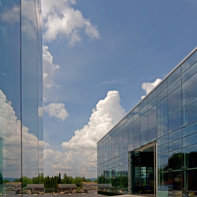 Blick zwischen zwei gläsernen Gebäudefassaden hindurch auf Bäume, darüber ein blauer Himmel mit weißen Wölkchen
