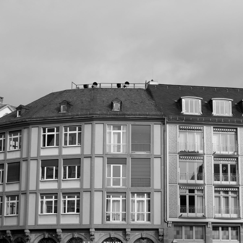 Schwarz-weiß-Foto eines vierstöckigen Wohn- und Geschäftshauses mit raumhohen Fenstern mit Oberlicht; die untere Etage ist nur im oberen Anschnitt zu sehen.