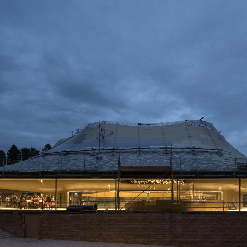 Pavillon mit geschwungenem Dach und großer umlaufender geschosshoher Verglasung aus gebogenem Glas bei Dunkelheit. Im erleuchteten Inneren ist eine Gruppe von Menschen links im Foyer zu sehen.