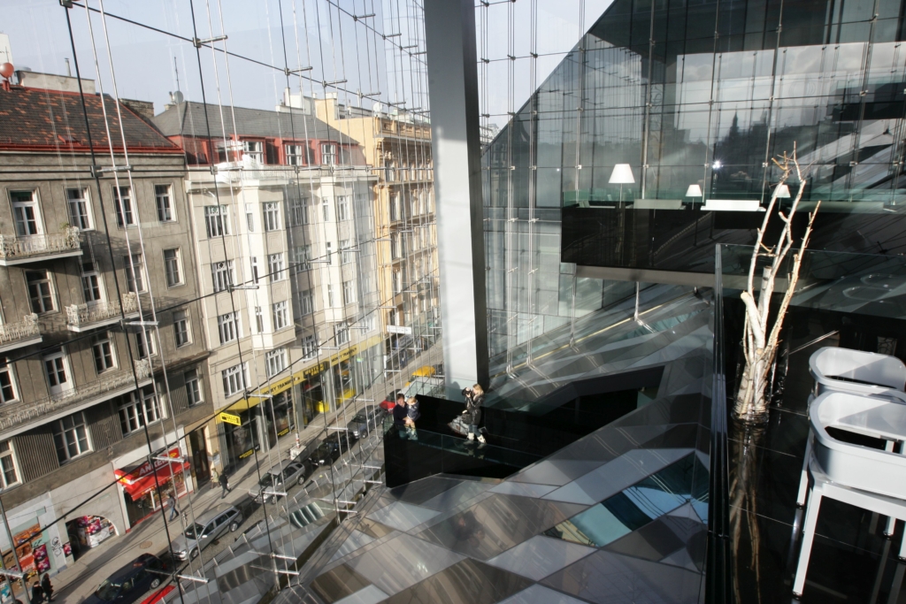 Block von oben in einen sehr großen, hohen Raum mit einer mehrgeschossigen Fensterfront links, die einen Blick auf die Straße darunter bietet. Der Fußboden besteht aus unterschiedlich geformten, blaugrauen geometrischen Platten. Rechts oben und hinten von schwarzen Glasbrüstungen gesäumte Galerien. Unten im Raum auf einer Art schwarzer Empore fotografiert eine Frau einen Mann mit einem Kind im Arm.