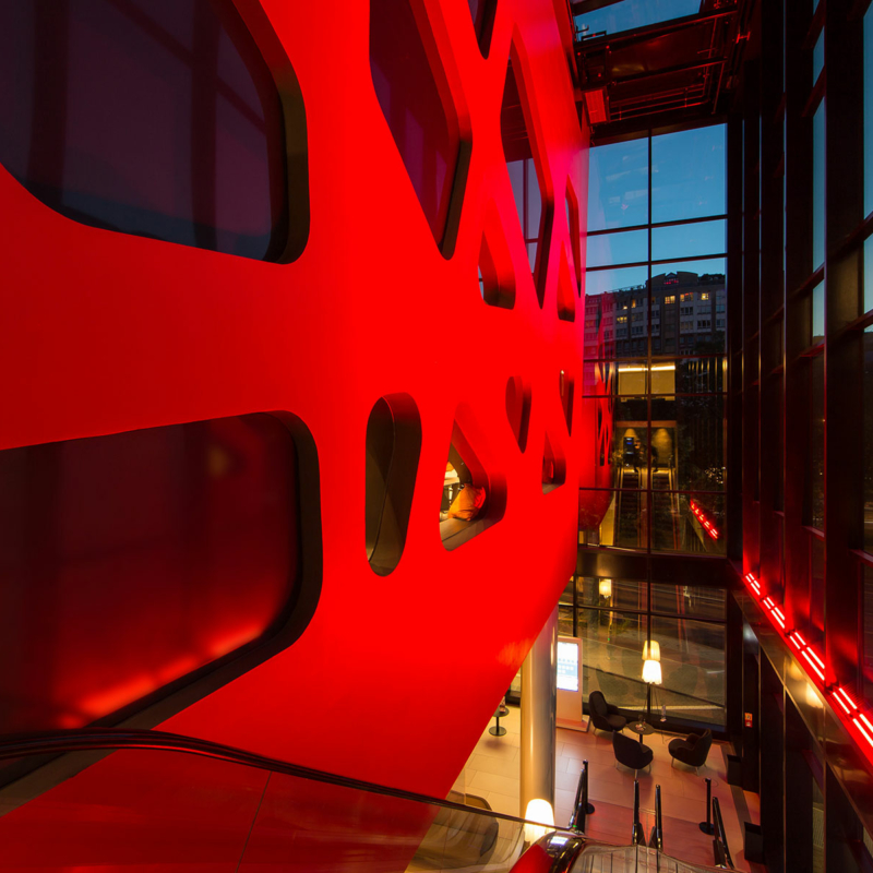 Blick zwischen zwei beleuchtete Fassadenhäute, die linke ist kräftig rot mit amorph geformten Fenstern. Durch den Spalt blickt man nach unten in eine Hotellobby mit Tischen und Stühlen, ebenfalls beleuchtet.
