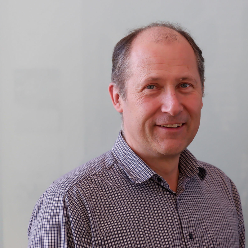 Thomas Kerscher, Geschäftsführer der Amberger Glas GmbH, blickt den Betrachter leicht zu seiner linken Seite abgewendet an. Er trägt ein am obersten Knopf offenes, kleinkariertes Hemd und steht vor einem hellen Hintergrund.