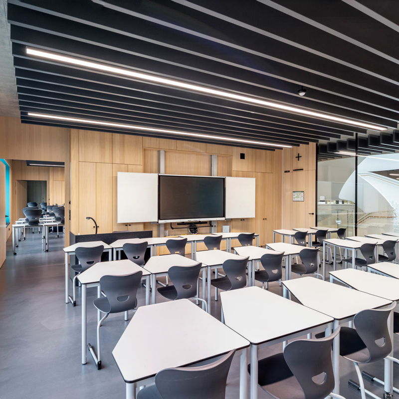 Klassenzimmer mit drei Tisch- und Stuhlreihen, links sichtbar ein sehr großes Fenster, rechts gläserne Trennwände zum Schulflur hin.
