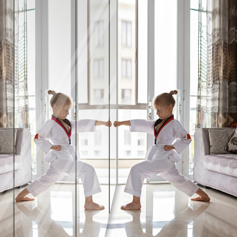 Ein kleines Mädchen steht in Karatehaltung vor einem bodentiefen Spiegelschrank, die rechte Faust ist nach vorne gestreckt und berührt fast den Spiegel. Rechts im Bild ein graues Sofa.